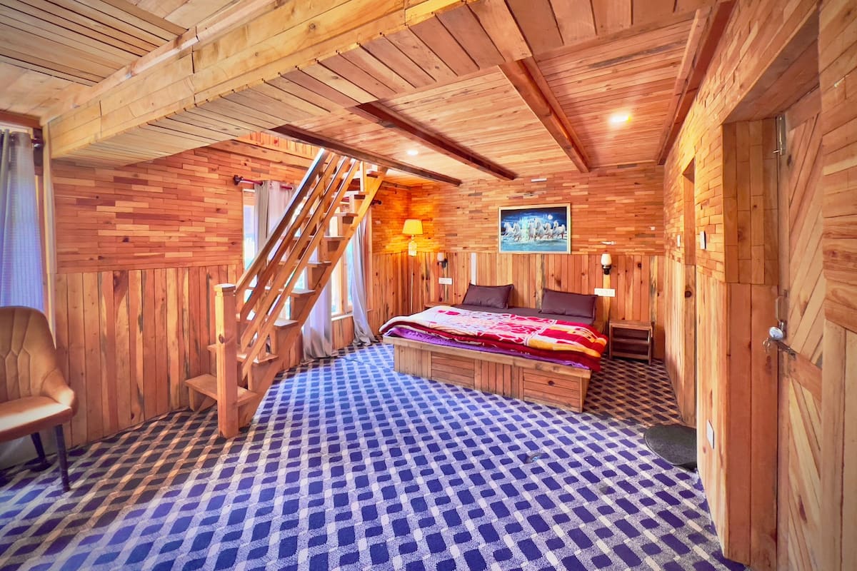 A wooden cabin bedroom with a checkered floor, a bed with a blanket, a chair, a wooden staircase, and a painting on the wall.