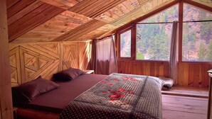 A wooden cabin bedroom with a bed, pillows, and a view of a forest through large windows. - Royal Star Kasol (Kasol)