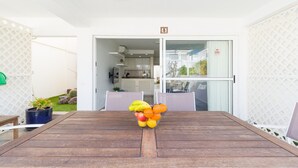 A wooden table with a fruit bowl in the center, a white door with a sign, and a view of a kitchen and garden through the door.