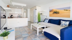 A modern living room with a blue sofa, white coffee table, and a kitchen area with white cabinets.