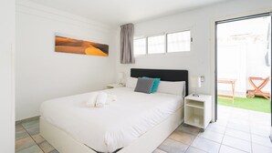 A bedroom with a large bed, a nightstand, a lamp, a painting, a window with curtains, and a view of a green outdoor area.