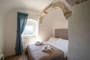 Habitación de hotel con cama, ventana con cortinas, arco de piedra y vistas al exterior.