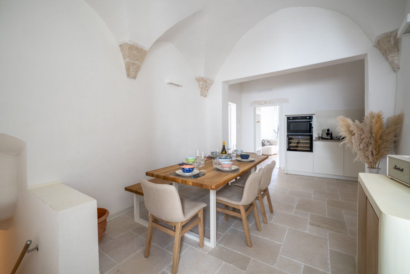 A dining area with a wooden table and chairs, a kitchen in the background, and a white interior.