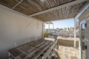 Une terrasse extérieure avec une table et des chaises, un espace abrité avec un toit en bambou, et une vue sur la mer et les bâtiments en arrière-plan.