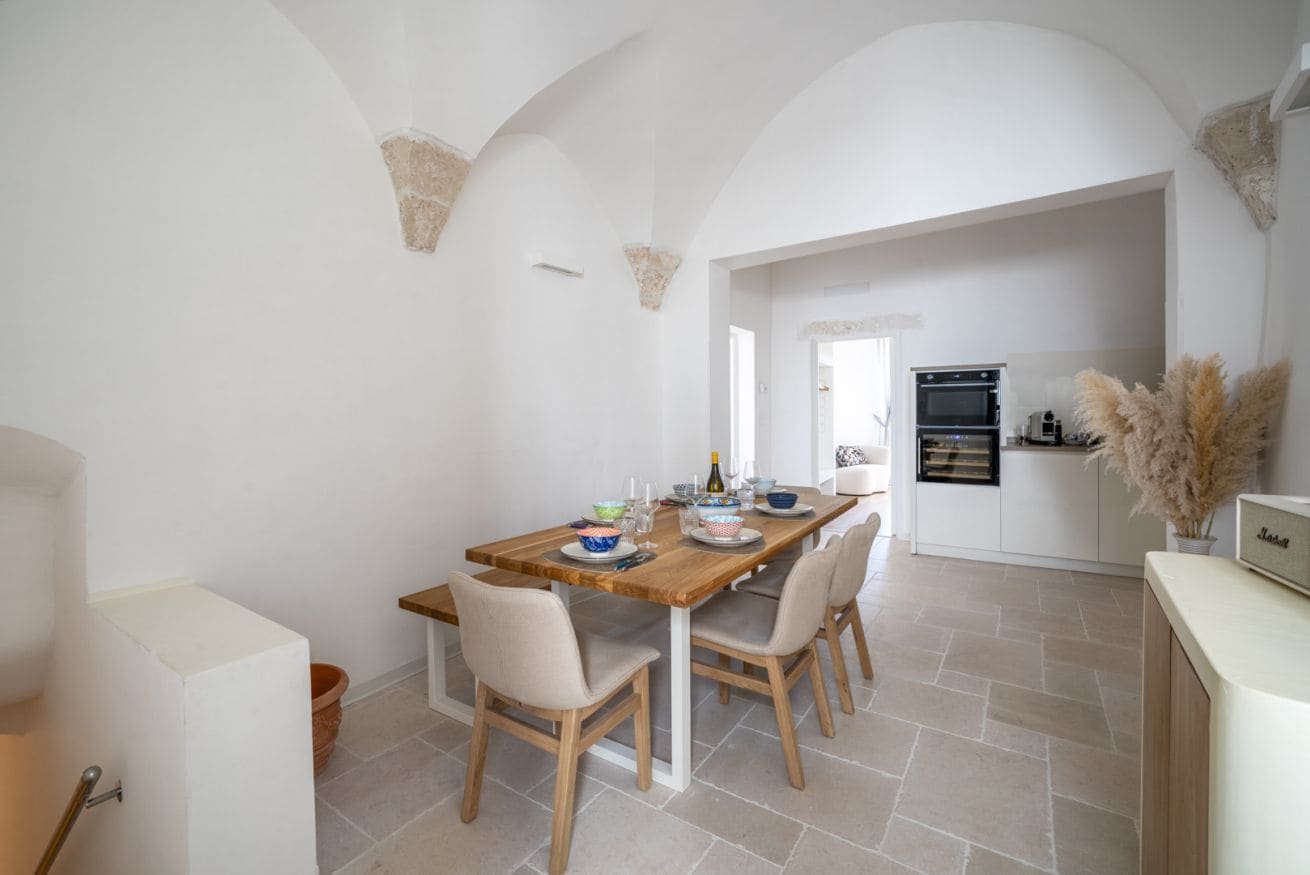 A dining area with a wooden table and chairs, a kitchen in the background, and a white interior.