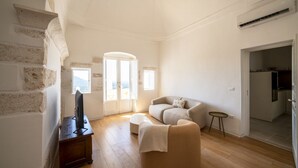 A living room with a sofa, ottoman, TV, and a small table. There is a door leading to another room and a window with a view. - Il Palazzotto: elegant historic residence in the heart of Ostuni with panoramic (Ostuni)