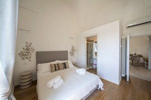 Une chambre à coucher moderne avec un grand lit, une table de chevet, une plante fixée au mur et une salle de bain visible à travers une porte ouverte.