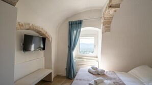 A bedroom with a TV, a bench, a bed with towels, a window with curtains, and a stone archway. - Il Palazzotto: elegant historic residence in the heart of Ostuni with panoramic (Ostuni)