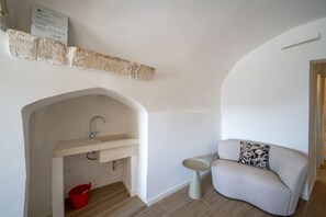 Una habitación pequeña con un lavabo integrado, un cubo rojo, una mesita redonda y un sofá con un cojín estampado.