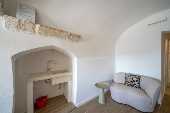 A compact room with a built-in sink, a red bucket, a small round table, and a sofa with a floral cushion.