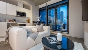 A modern living room with a sofa, a coffee table, a kitchen area, and a large window with a city view.
