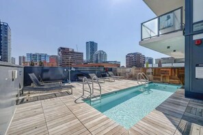Une piscine sur le toit, avec une terrasse en bois, des échelles métalliques et une vue sur les toits de la ville en arrière-plan.