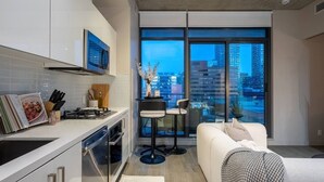 A modern kitchen with a dining area, a sofa, and a large window with a city view.