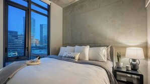A modern hotel room with a large bed, a nightstand with a lamp, a potted plant, and a city view through large windows.