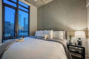 Quarto de hotel moderno com uma cama grande, mesa de cabeceira com abajur, planta em vaso e vista para a cidade através de grandes janelas.