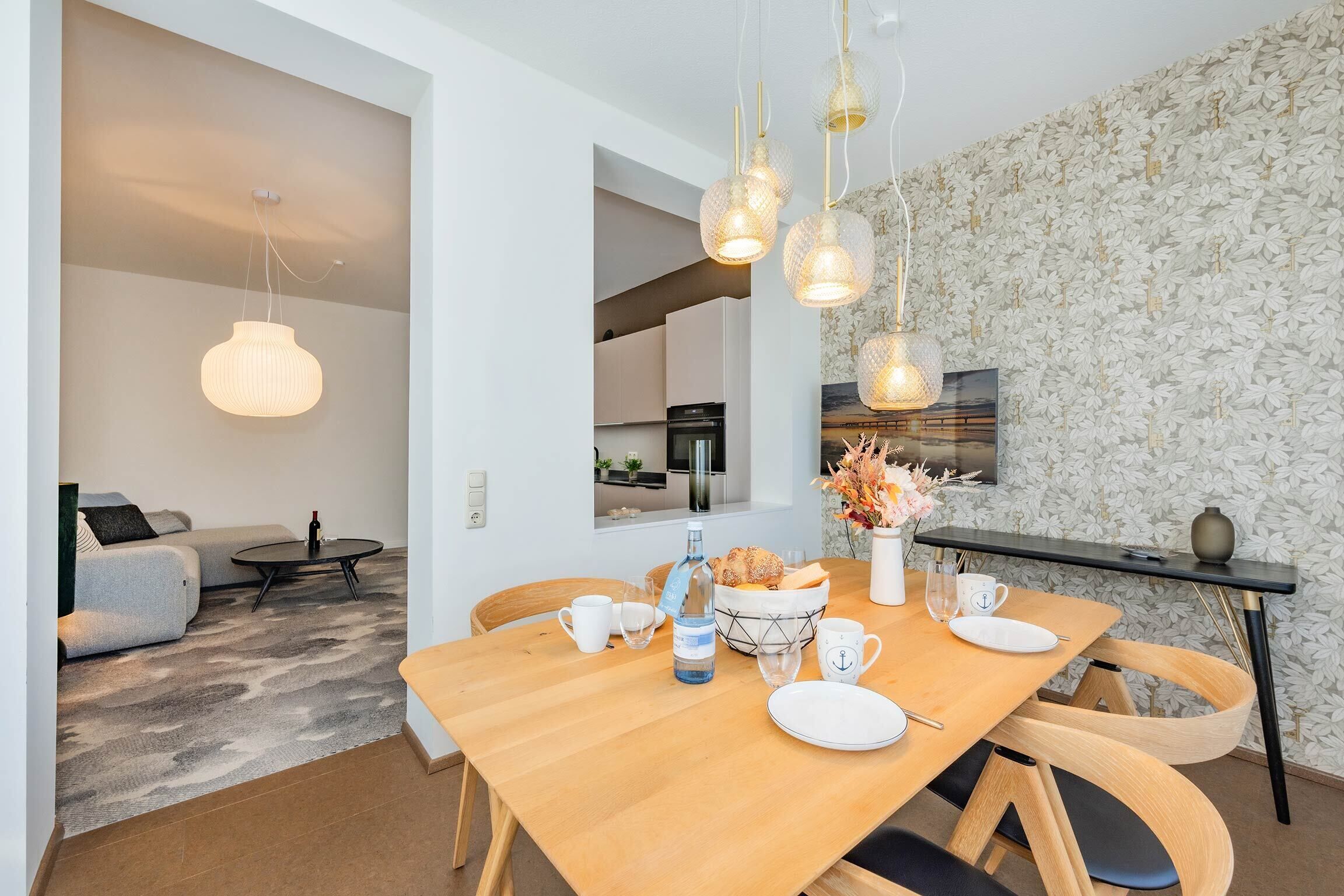 A dining area with a wooden table set for four, a vase with flowers, and a kitchen in the background.