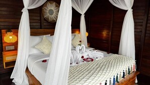 A four-poster bed with white linens, a heart-shaped towel, and rose petals. - Eco Oasis Wooden Villa (Tegallalang)
