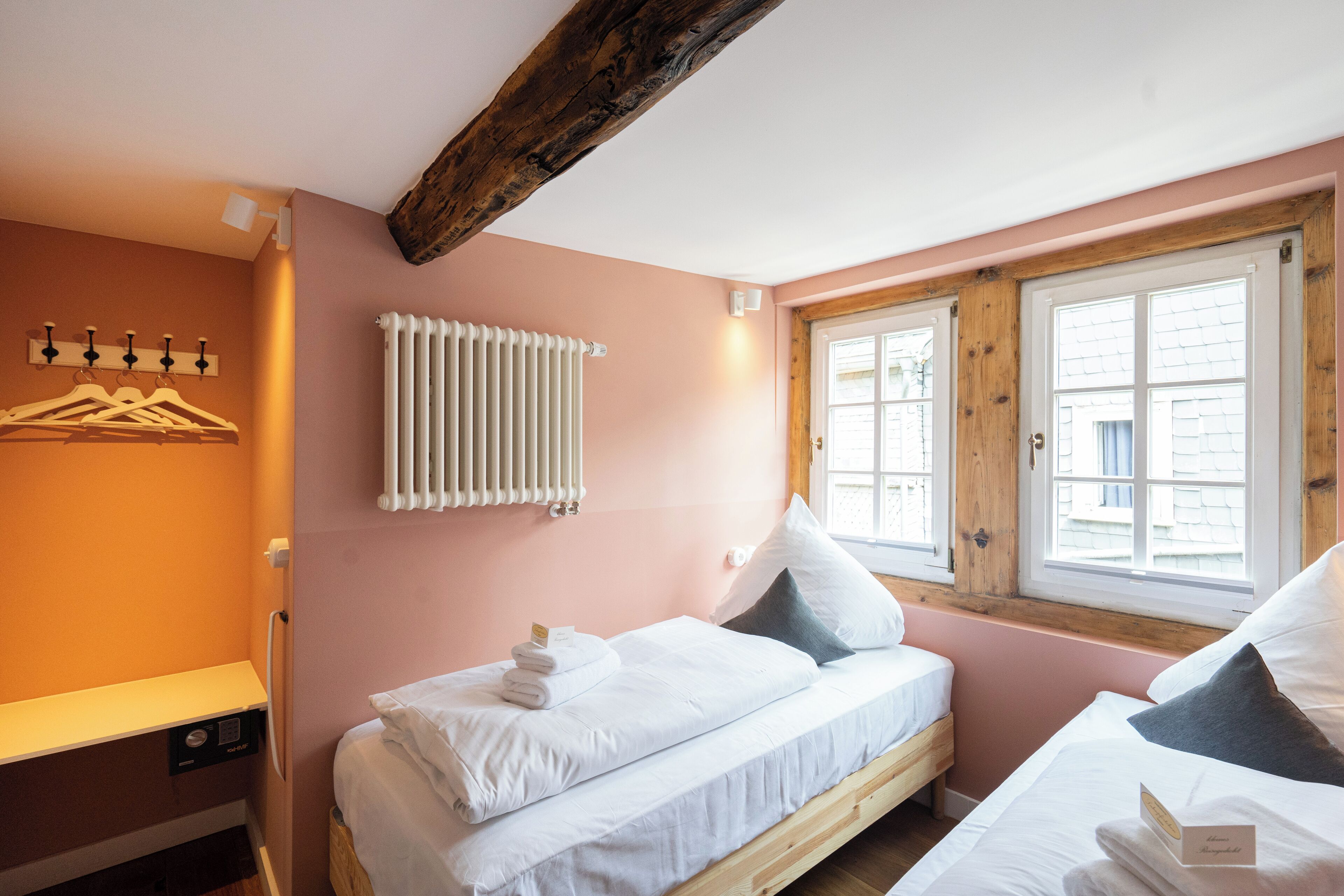 A bedroom with two beds, a radiator, a window with wooden shutters, and a wall-mounted light fixture.