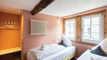 A bedroom with two beds, a radiator, a window with wooden shutters, and a wall-mounted light fixture.