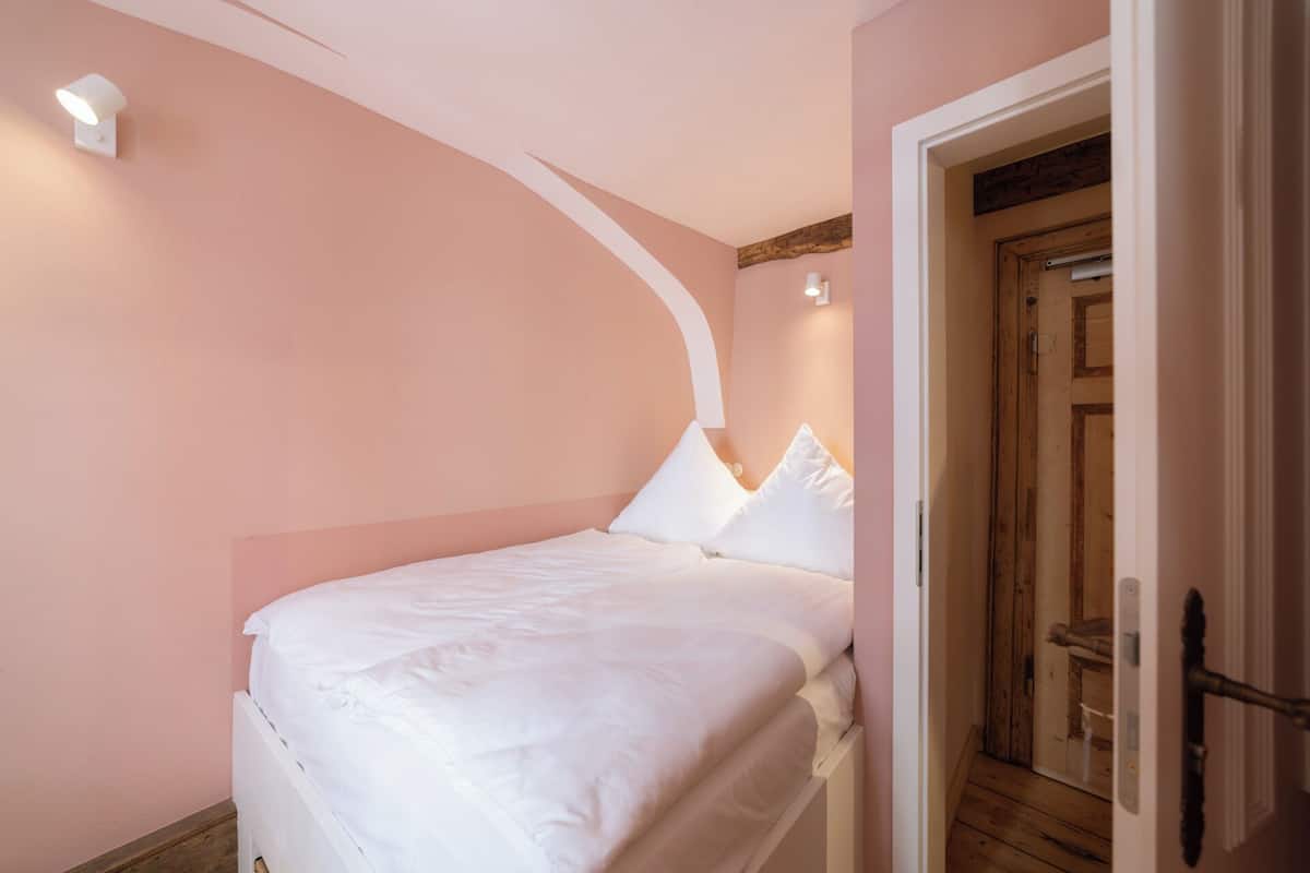 Une chambre avec un lit simple, du linge de lit blanc, un mur incurvé et une porte en bois.