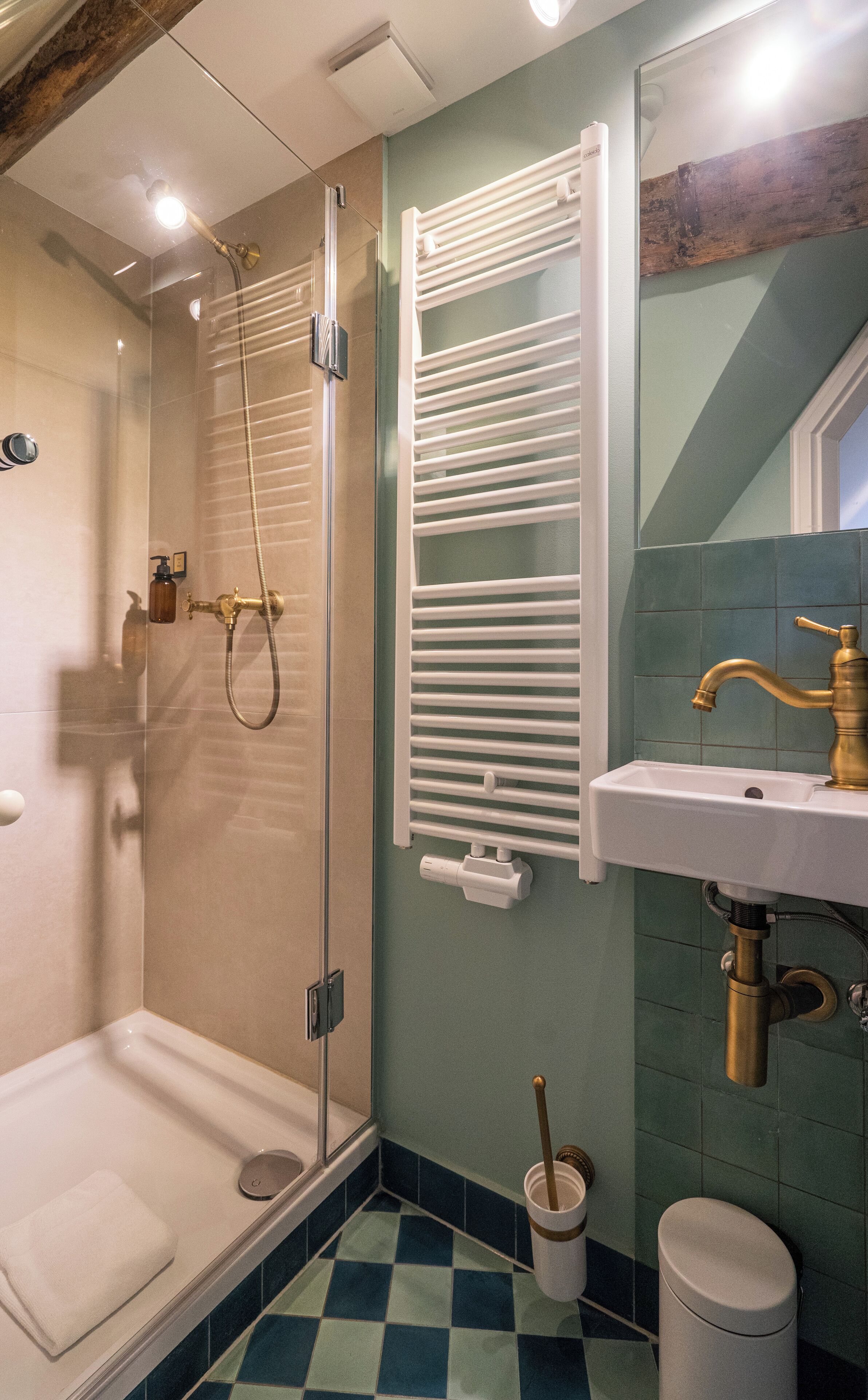 A bathroom with a glass shower enclosure, a white towel rack, a sink with a gold faucet, and a trash bin.