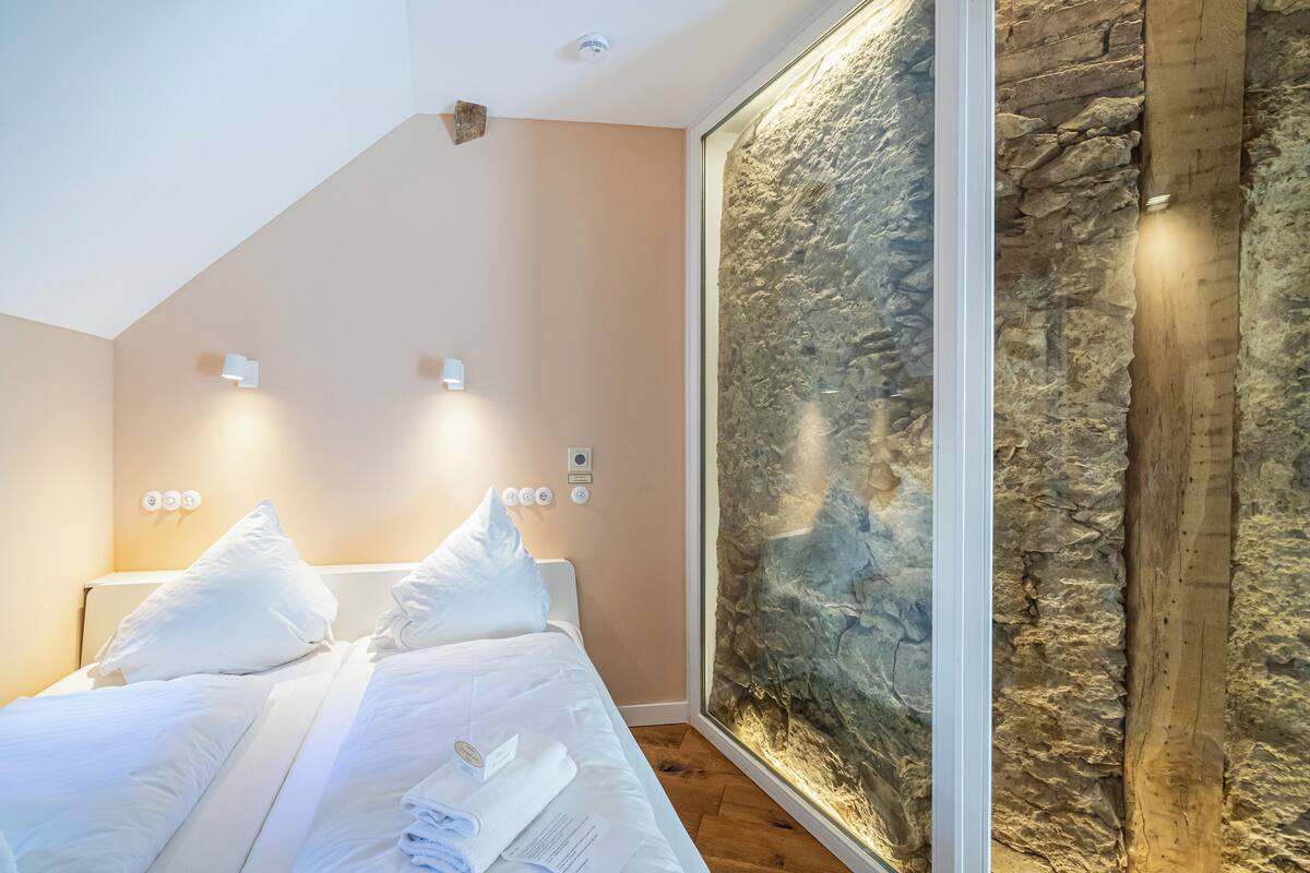Un lit bien fait, avec du linge de lit blanc et des oreillers, un mur en pierre décoratif et un luminaire moderne au plafond.