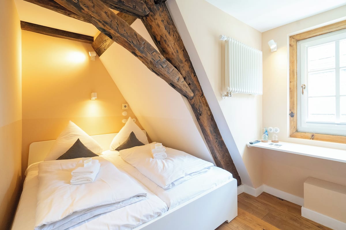 Un lit bien fait, avec des oreillers et une serviette pliée, une structure en poutres apparentes, une fenêtre donnant sur un paysage agréable et un radiateur fixé au mur.