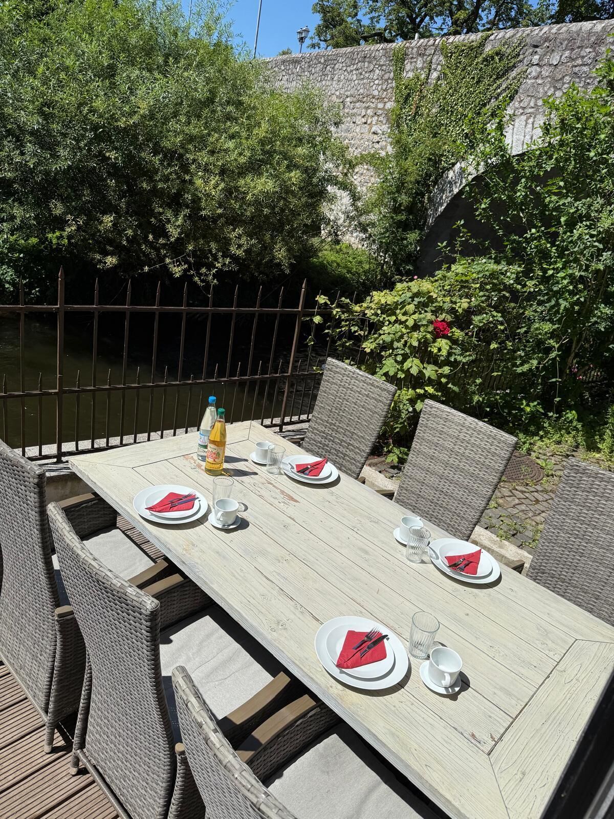 An outdoor dining area with a table set for four, surrounded by wicker chairs, near a stone wall and greenery.
