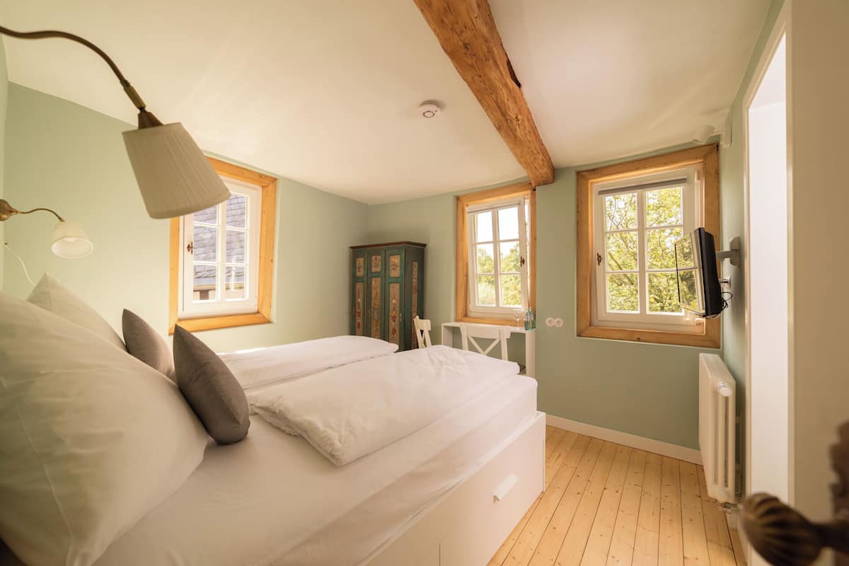 Une chambre à coucher avec un lit, deux fenêtres, une poutre en bois au plafond, une armoire et un radiateur.