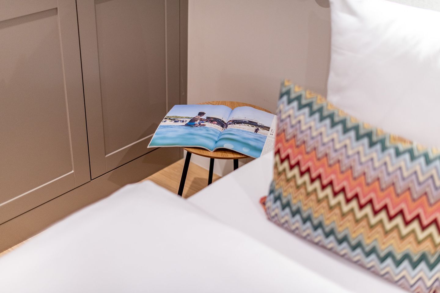 A bed with a white sheet, a colorful zigzag pillow, and an open magazine on a small wooden stool.