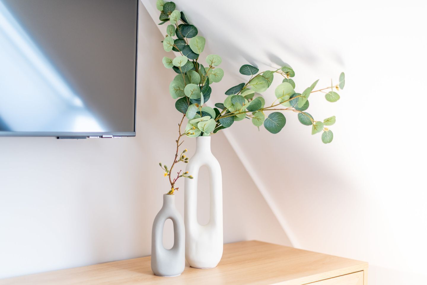 Two white vases with a plant on a wooden surface against a wall.