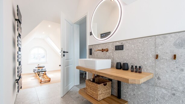 A modern bathroom with a white sink, a mirror with a light, and a wooden shelf with toiletries. - F'27 Living, Whg. 06 - F27 Living (Sylt)