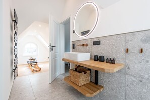 Un baño moderno con un lavamanos blanco, un espejo con luz y un estante de madera con artículos de aseo.
