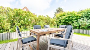 Een houten terras buiten, met een tafel en stoelen, omringd door weelderige begroeiing en een houten hek.