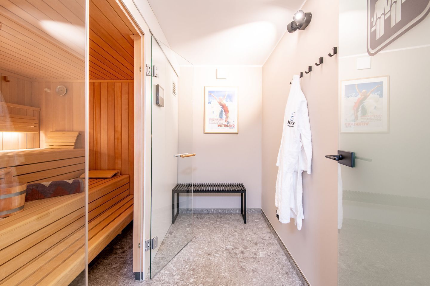 Ein Saunaraum mit Holzbänken und einer Glastür zu einem Duschbereich mit Sitzbank und Handtüchern an der Wand.
