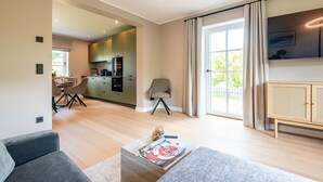 Een moderne woonkamer met een bank, een salontafel met tijdschriften, een eethoek met stoelen, een keuken en een groot raam met gordijnen.