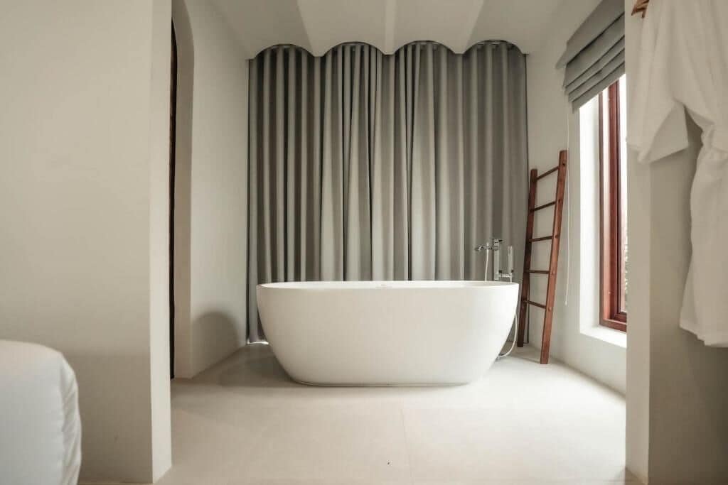 A modern bathroom with a freestanding bathtub, a wooden ladder, and a window with curtains.