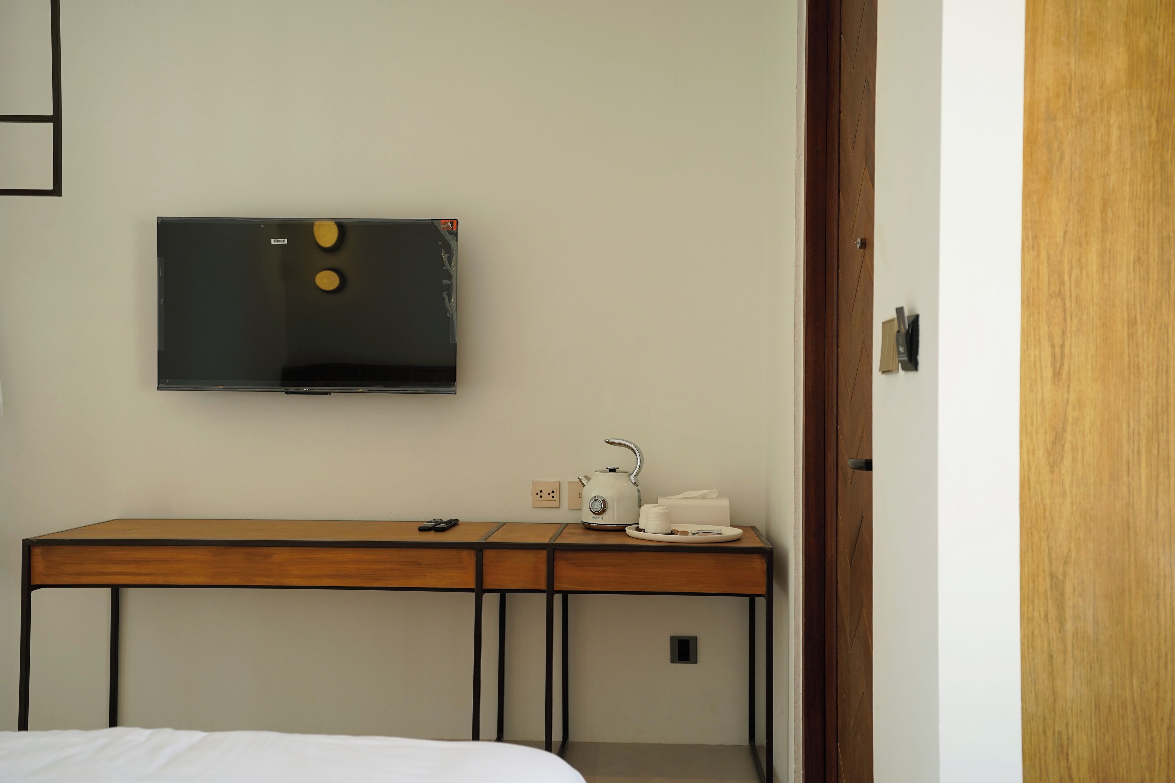 A hotel room with a wooden desk, a flat-screen TV, a kettle, and a cup.