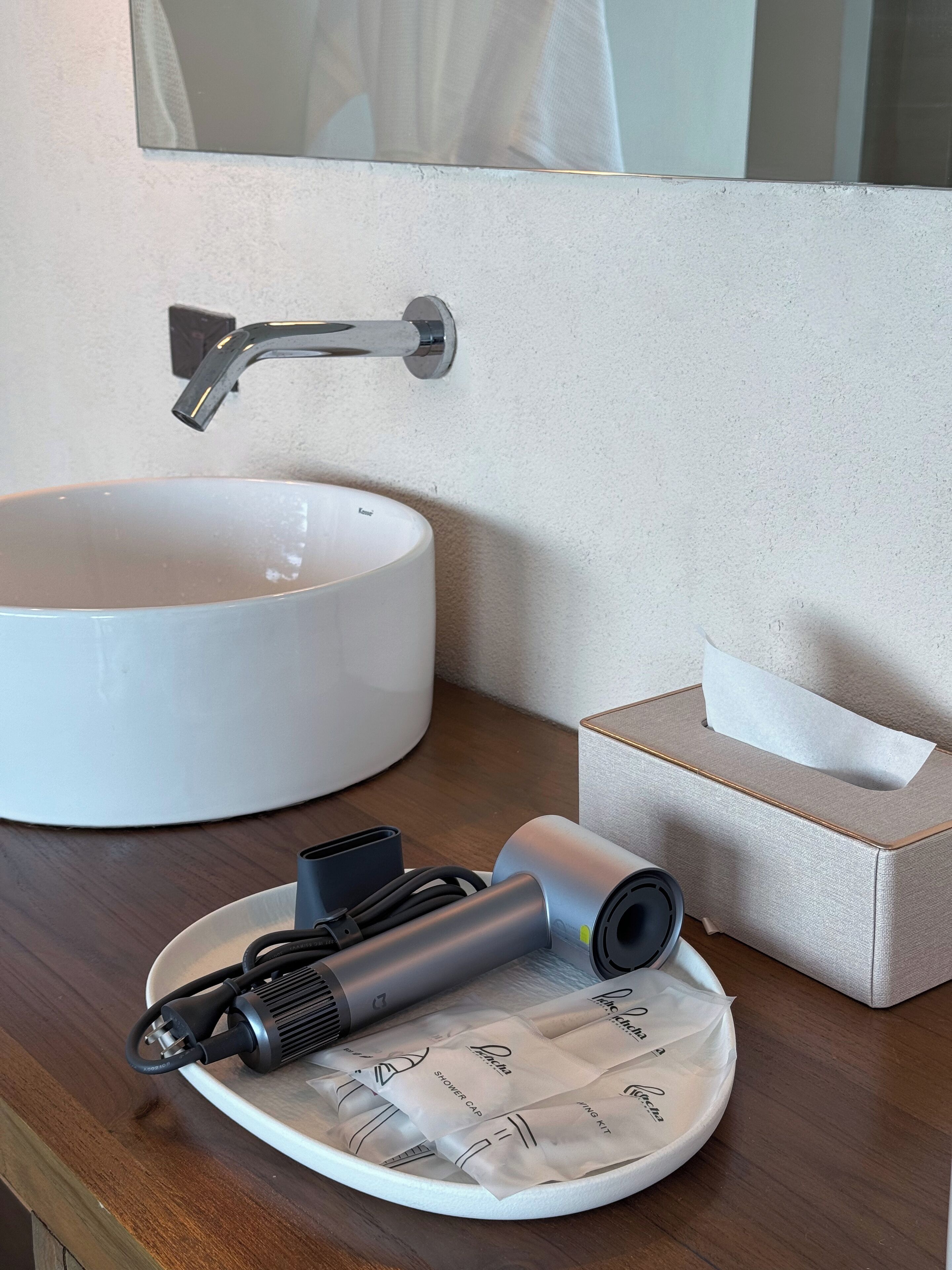 A bathroom with a round white sink, a modern faucet, a tissue box, and a hairdryer on a wooden countertop.