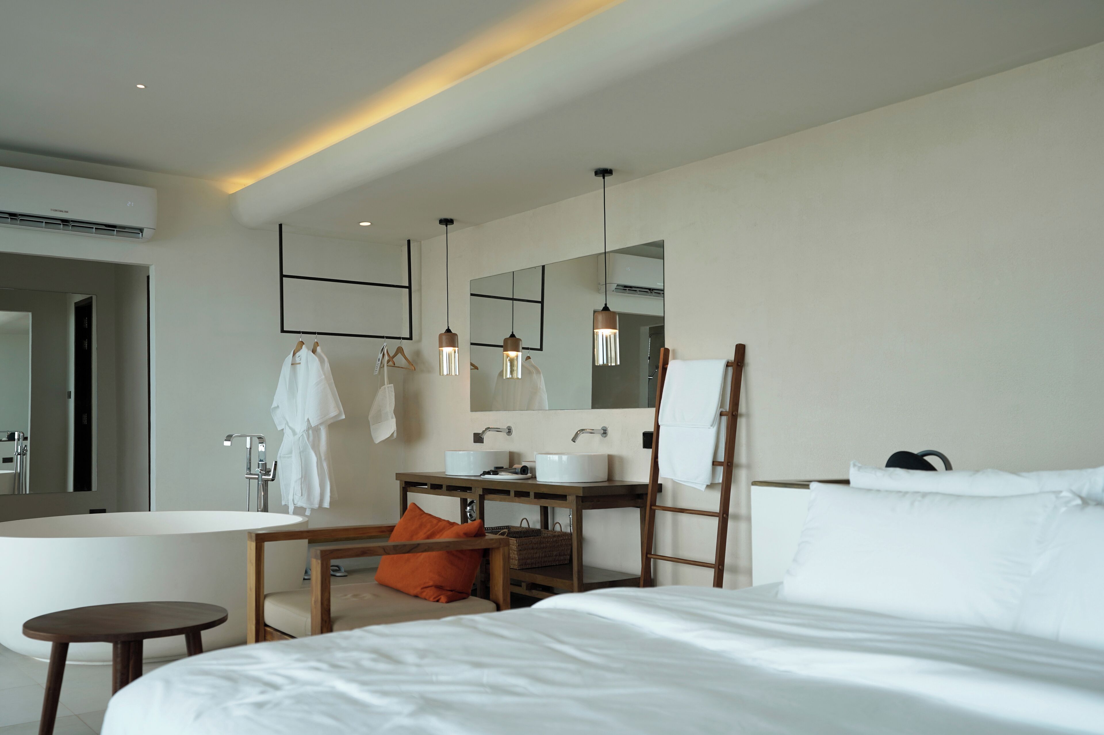 A modern hotel room with a large bed, a wooden bedside table, a small round table, a bathtub, a vanity with a mirror, and a large mirror above the vanity.