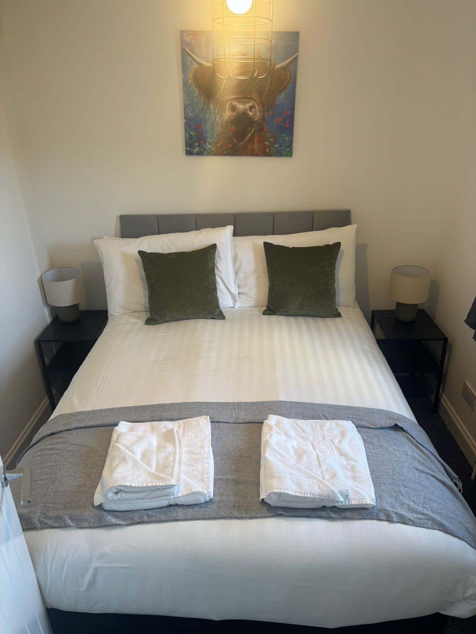 Ein ordentlich bezogenes Bett mit weißen Leinentüchern, zwei Kissen und zwei gefalteten Handtüchern. Darüber hängt ein Gemälde, und auf jeder Seite des Bettes stehen zwei Nachttischlampen.