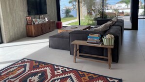 En moderne stue med en stor sofa, et sofabord og et fjernsyn monteret på en betonvæg. Store vinduer giver udsigt til det fri.