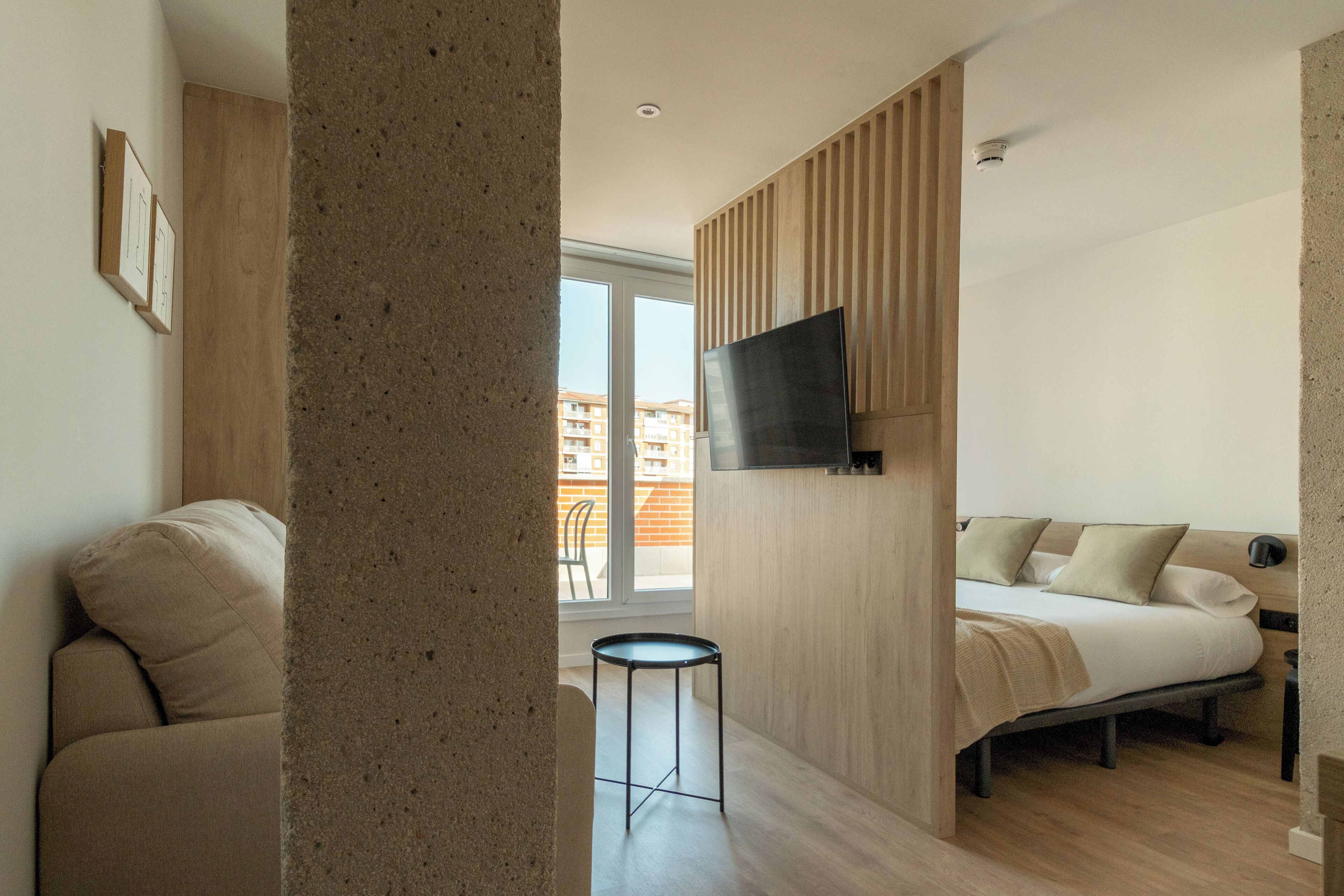 Een moderne hotelkamer met een bed, een aan de muur bevestigde televisie, een klein tafeltje en uitzicht op het balkon.