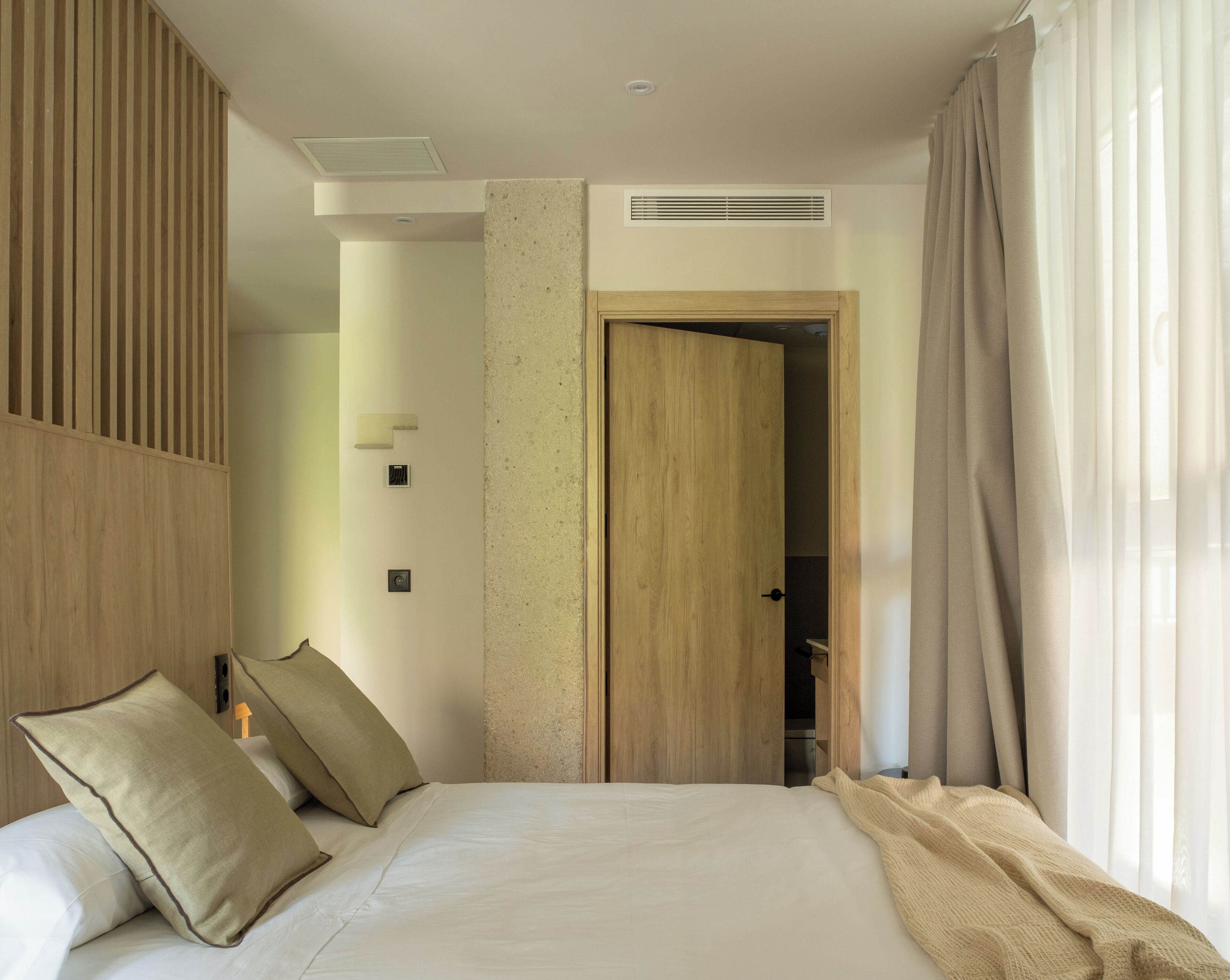 Ett modernt hotellrum med en stor säng, träpanel på väggarna, en trädörr och utsikt över korridoren.