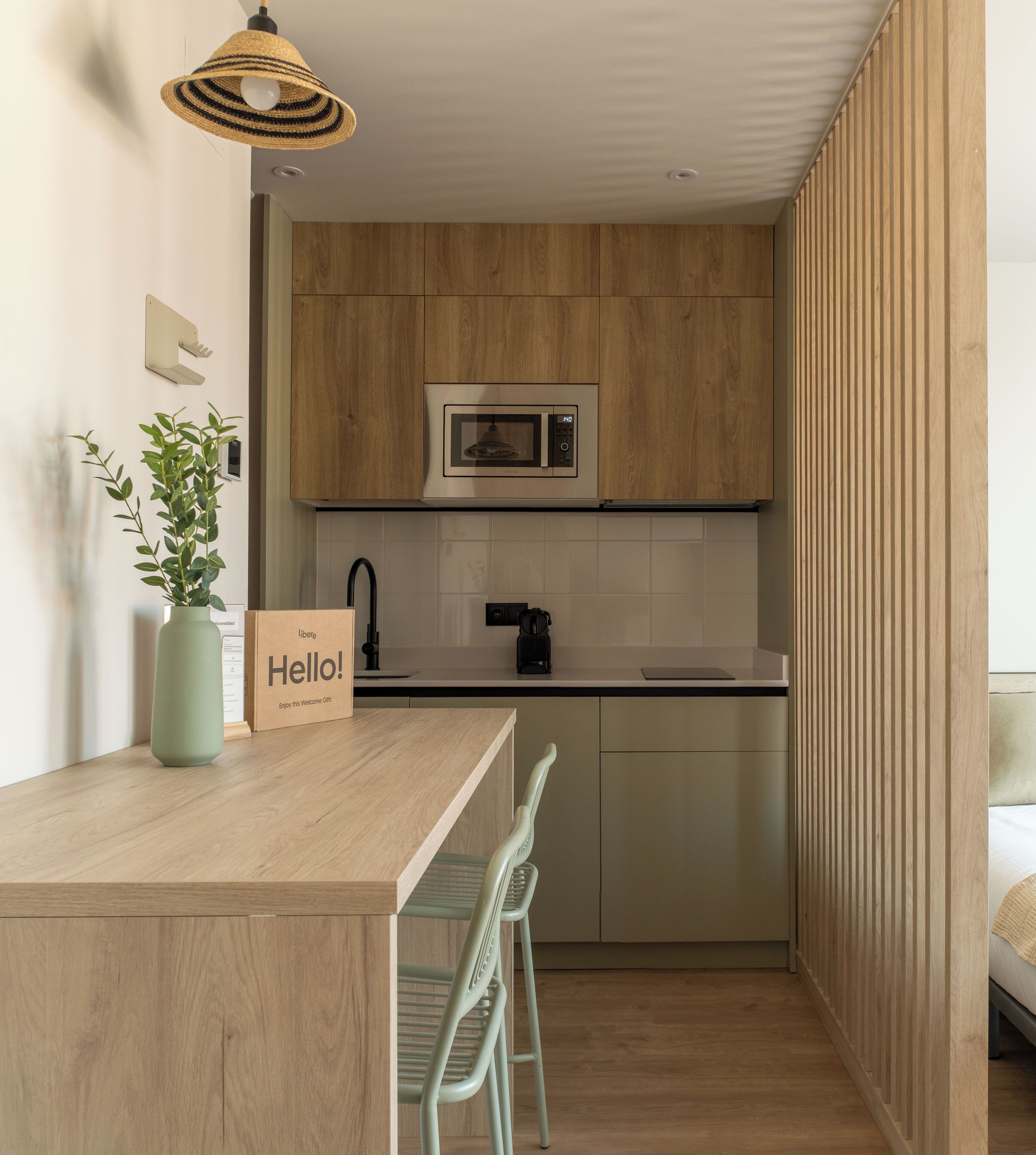 Een moderne keuken met houten kastjes, een ingebouwde magnetron, een spoelbak en een klein eethoekje met een stoel.