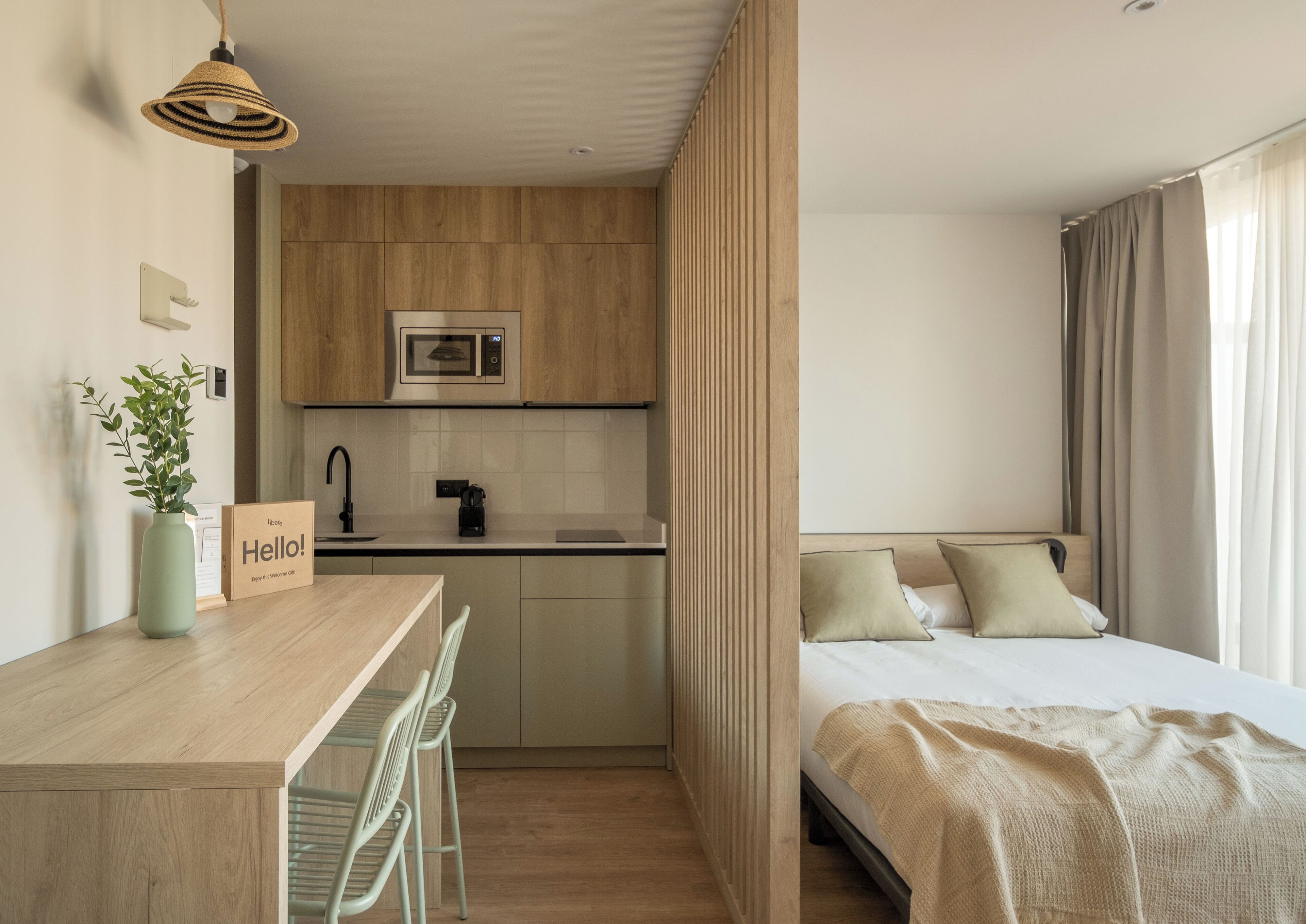 Habitación de hotel compacta con kitchenette, una cama con manta color beige y vistas al exterior a través de la ventana.