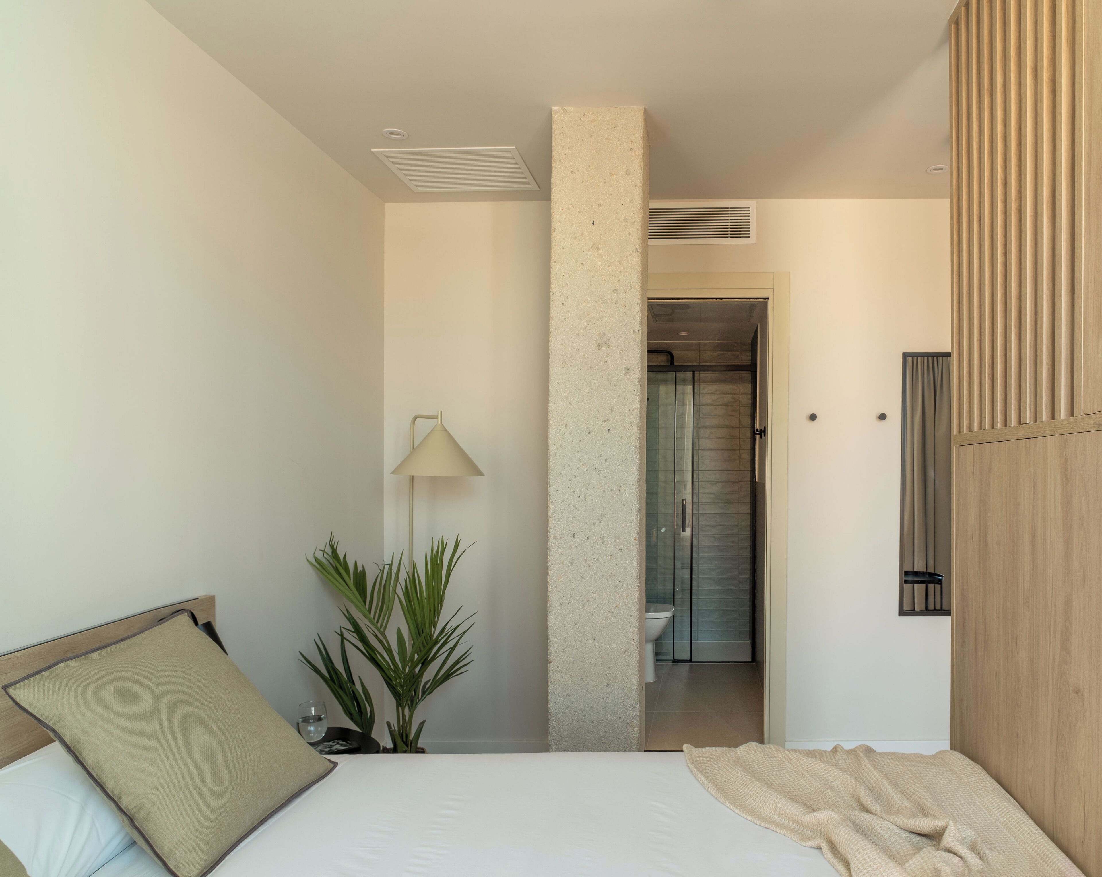 A modern hotel room with a bed, a bedside lamp, a potted plant, and a view into the bathroom.
