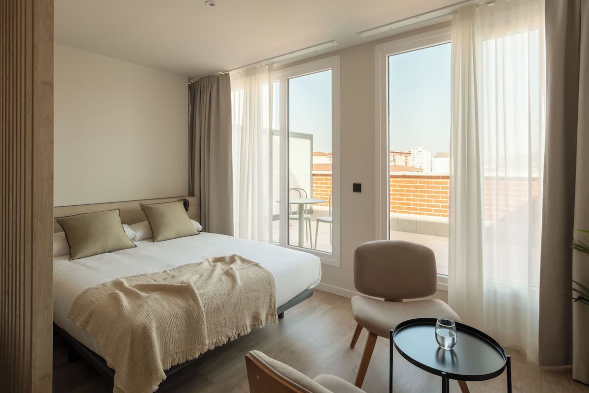 A hotel room with a bed, a chair, a small table, and large windows.