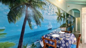 A dining area with a mural of a tropical beach scene, a table with a floral tablecloth, and white cabinets. - Terrazza sul Mare - by BelcantoVillas (Lido Di Camaiore)