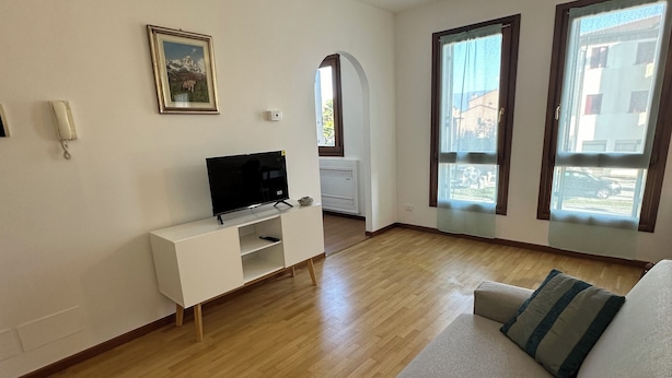 A modern living room with a white sofa, a flat-screen TV on a white stand, and a large window with curtains.